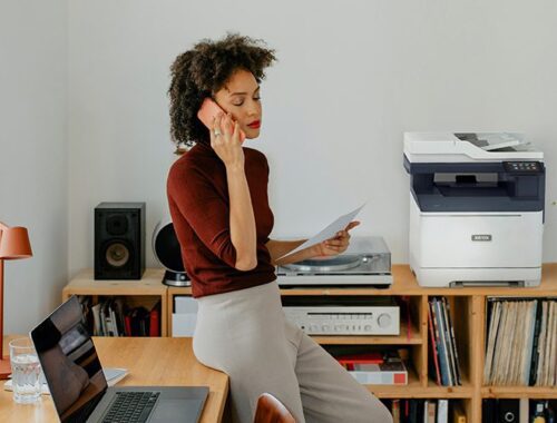 Una mujer sentada en un escritorio teniendo una llamada junto a una impresora Xerox® C325