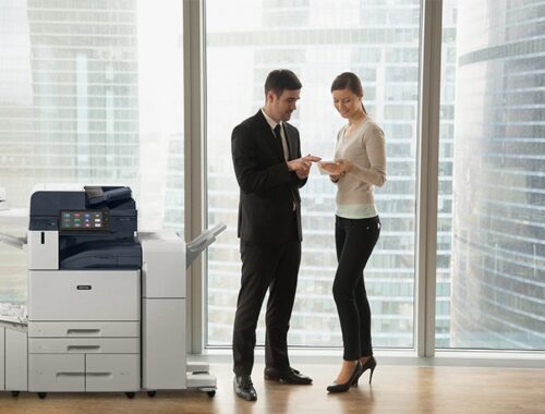 Dos personas en una oficina vestidas de negocios discutiendo documentos junto a una impresora multifunción Xerox® AltaLink® serie B8200